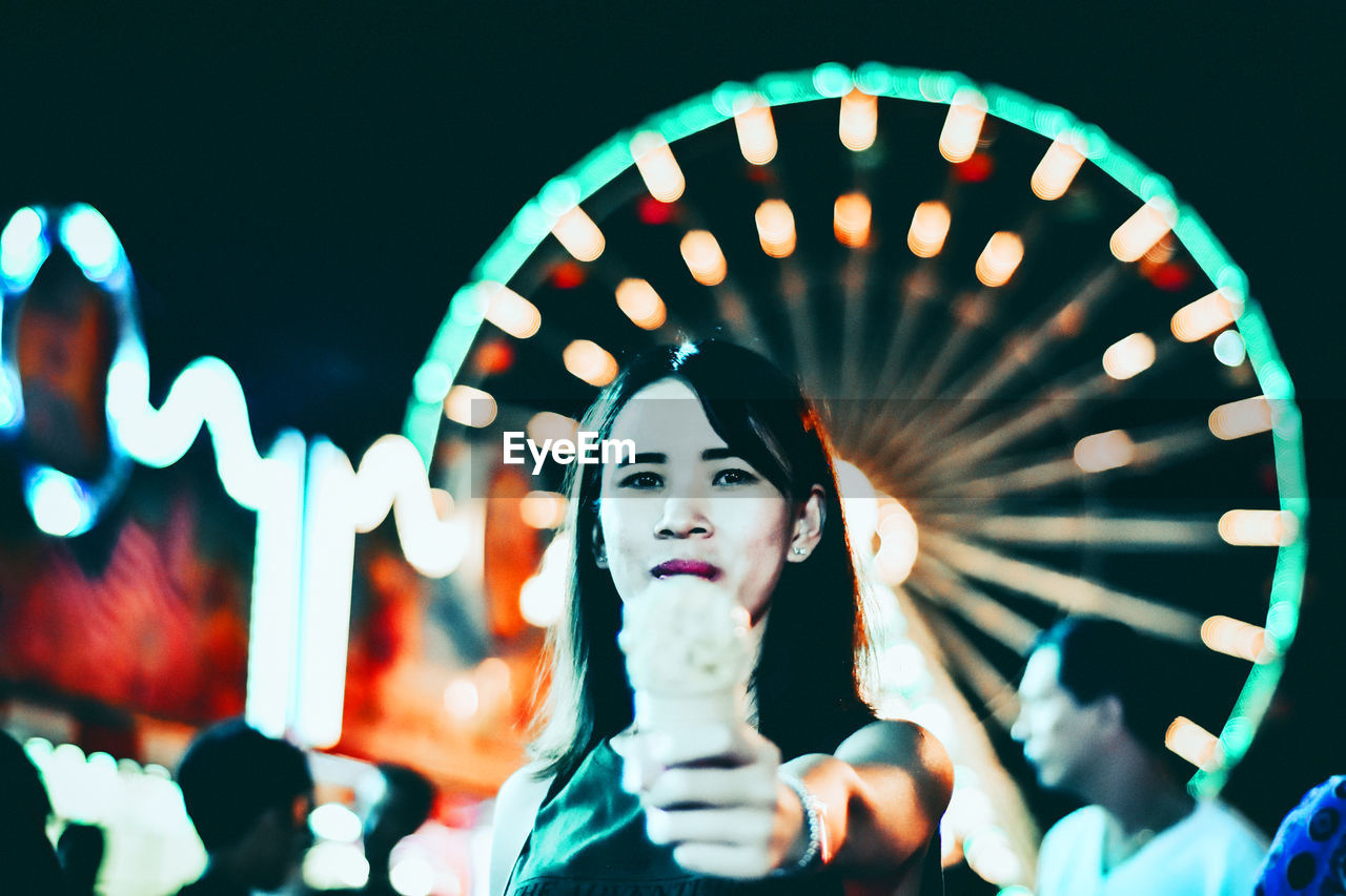 Portrait of smiling young woman standing holding ice cream in city at night