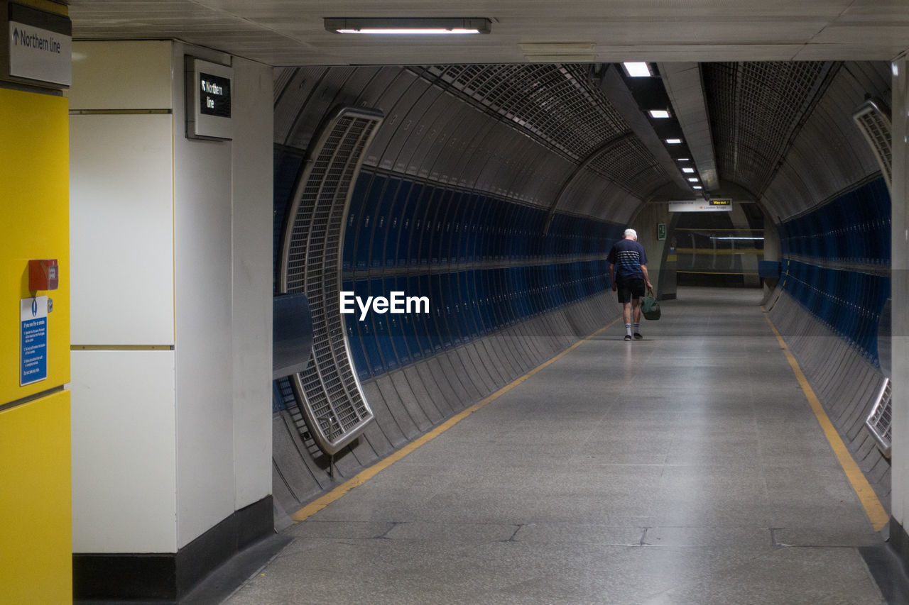 REAR VIEW OF MAN WALKING IN ILLUMINATED UNDERGROUND WALKWAY