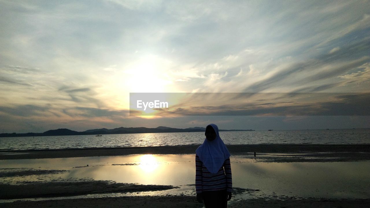 Woman standing at beach against sky during sunset