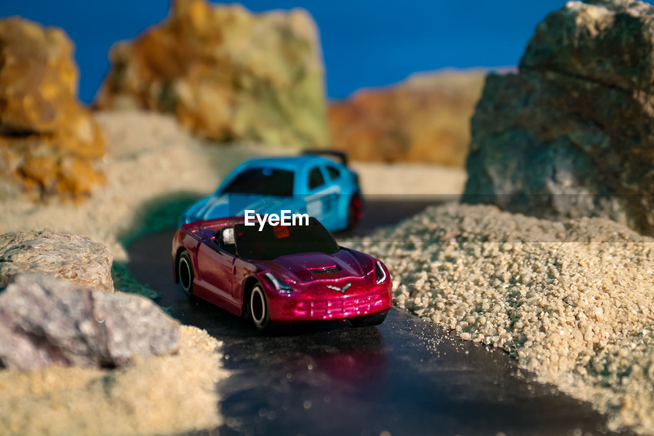 car, vehicle, rock, land vehicle, nature, transportation, mode of transportation, motor vehicle, travel, blue, land, sky, no people, selective focus, sunlight, water, red, day, outdoors, travel destinations, adventure, screenshot, focus on foreground, sunny, toy car