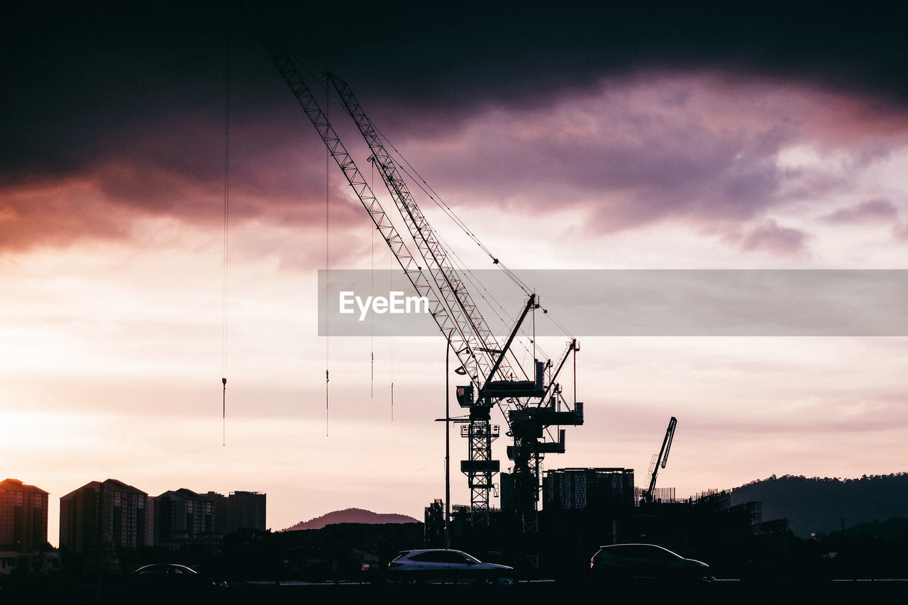 Silhouette tower cranes on construction site in city against sky at sunset