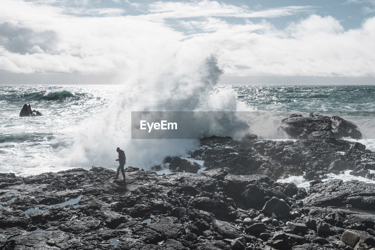 Full length of hiker against waves splashing on rocks at beach