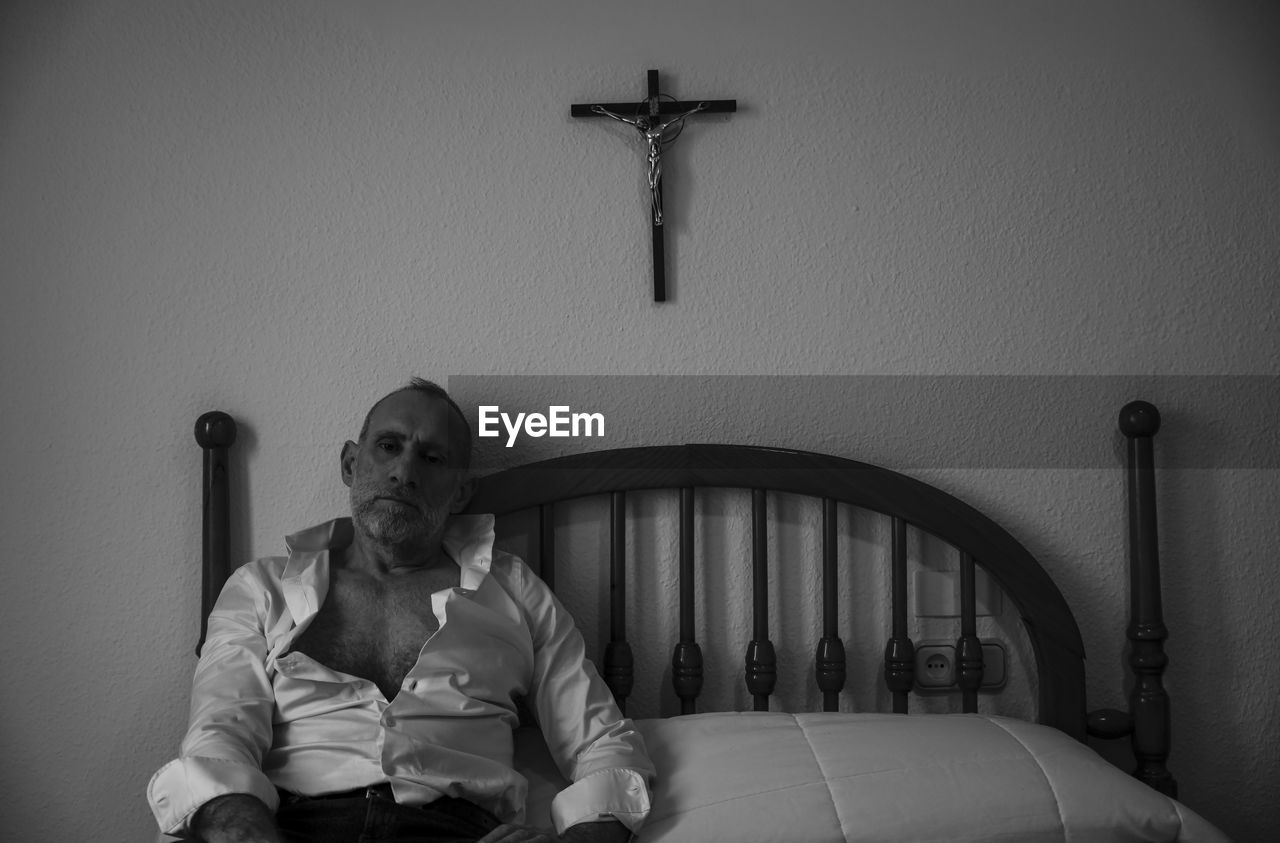 Monochrome of adult man in white shirt in room lying on bed with religion cross on wall