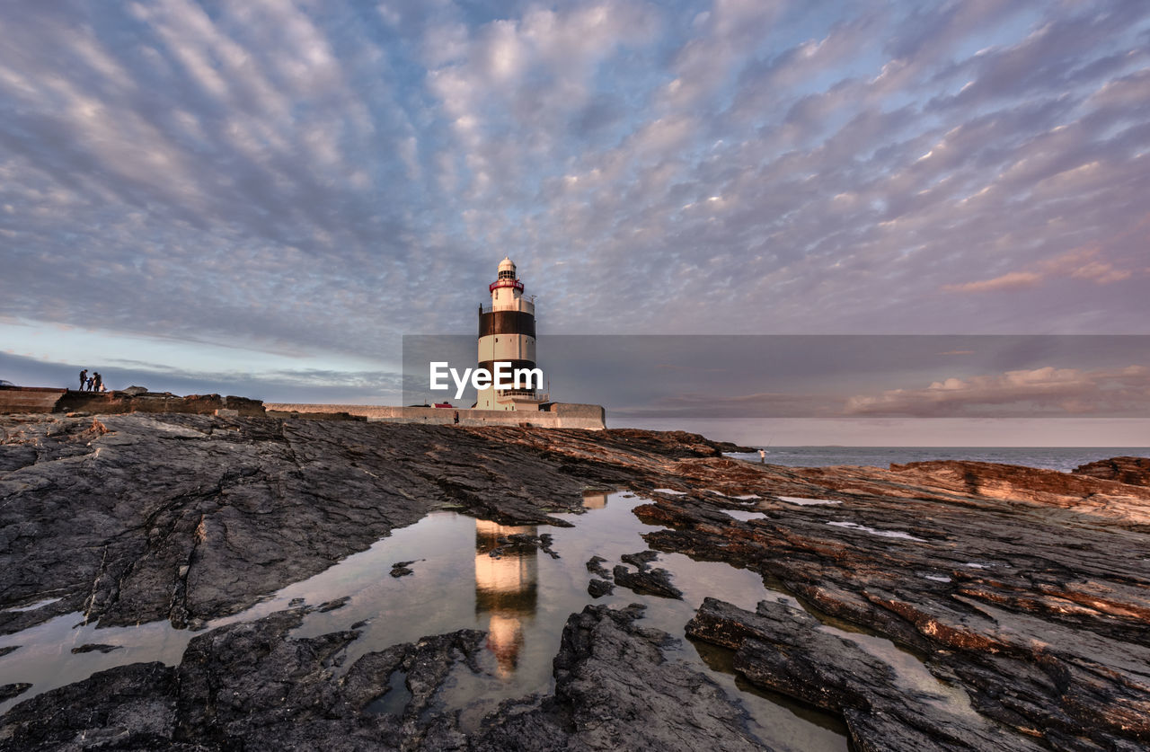 Lighthouse by sea against cloudy sky during sunset