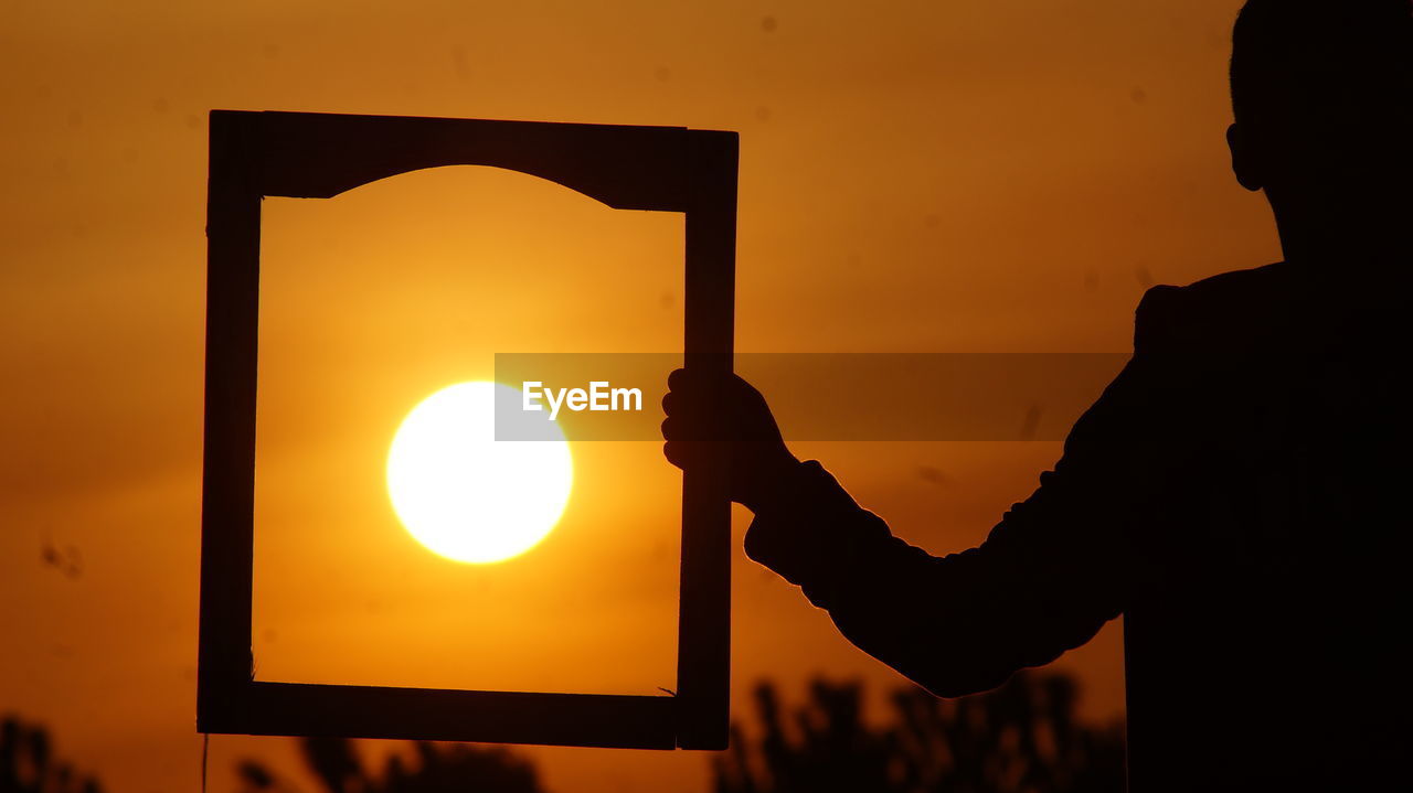 Silhouette man holding picture frame in front of sun during sunset