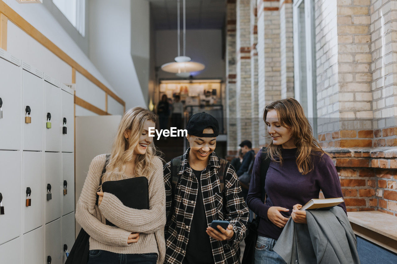 Smiling man sharing smart phone to female friends while walking in corridor