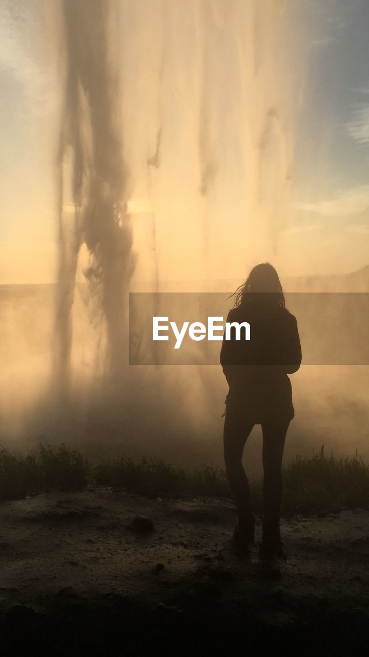 Rear view silhouette of woman standing by hot spring against sky during sunset