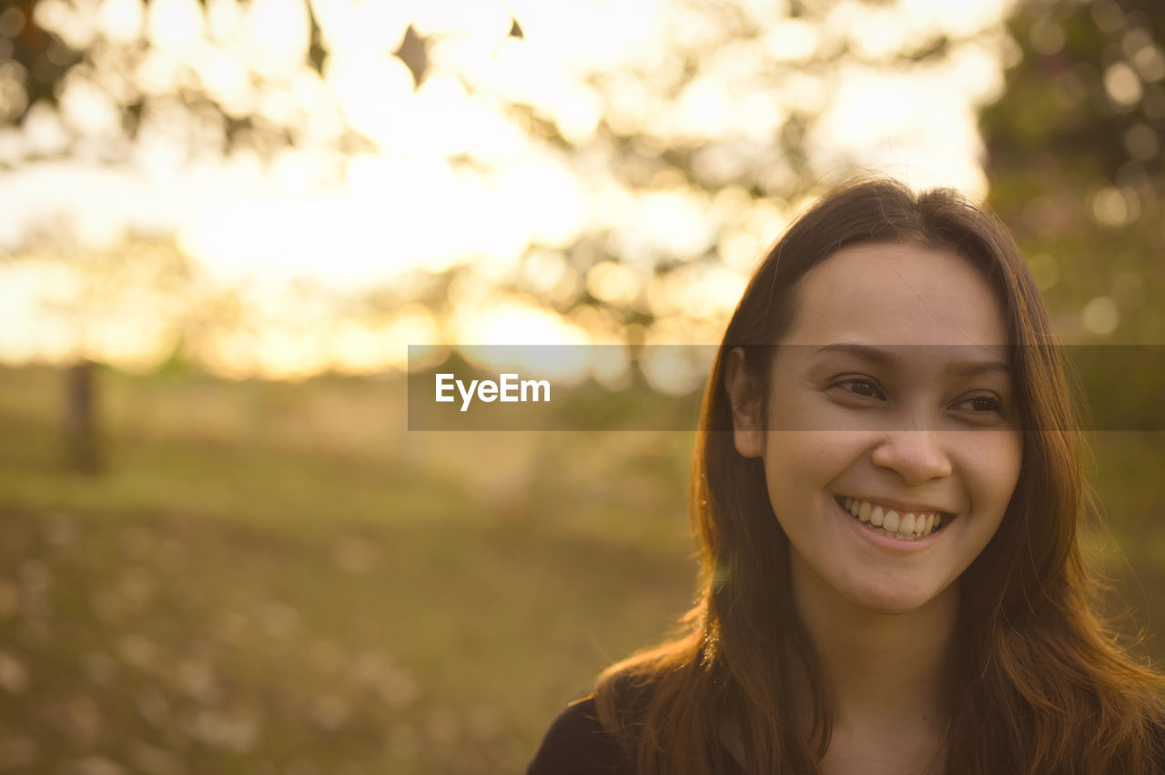 smiling, portrait, happiness, one person, long hair, emotion, headshot, adult, hairstyle, sunlight, young adult, women, portrait photography, female, looking at camera, nature, cheerful, teeth, smile, brown hair, front view, enjoyment, autumn, focus on foreground, person, back lit, copy space, outdoors, tree, positive emotion, sky, sunset, human face, casual clothing, lifestyles, beauty in nature, leisure activity, day, environment, relaxation, landscape, joy, carefree, land, plant, rural scene, lens flare, yellow, tranquility