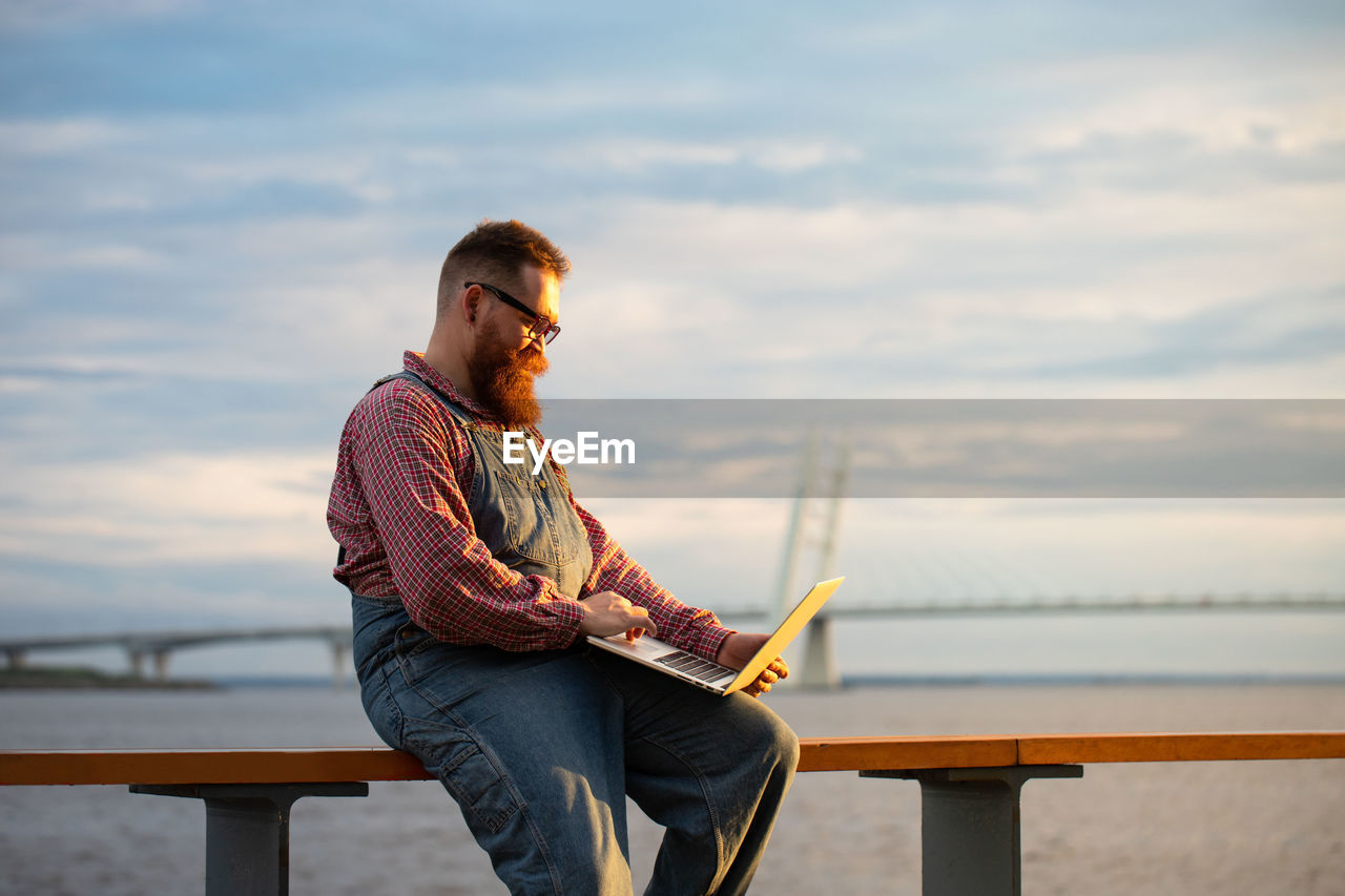 Bearded hipster man freelancer using laptop,  writing message, chatting, shopping at embankment 