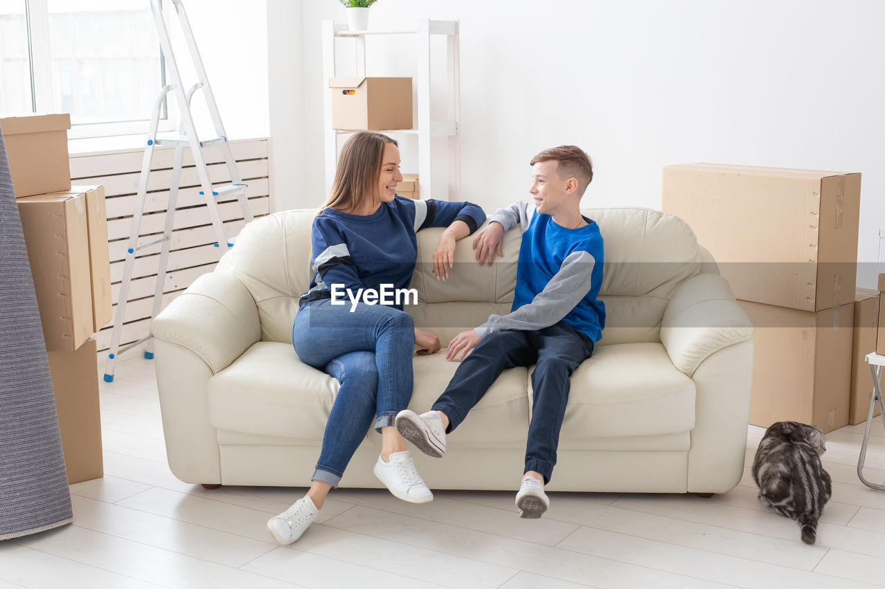 FULL LENGTH OF YOUNG WOMAN SITTING ON SOFA AT HOME