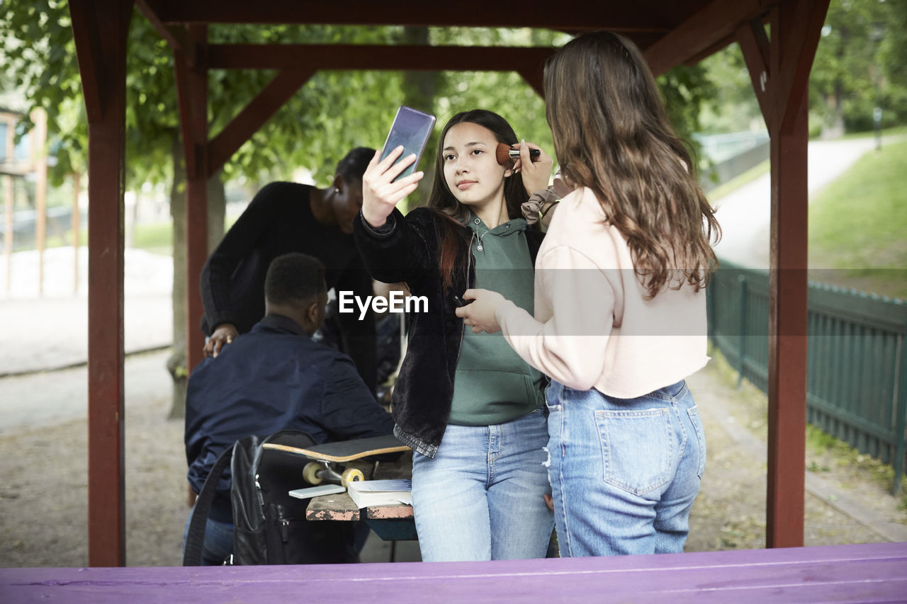 Teenage girl applying make-up to female friend taking selfie