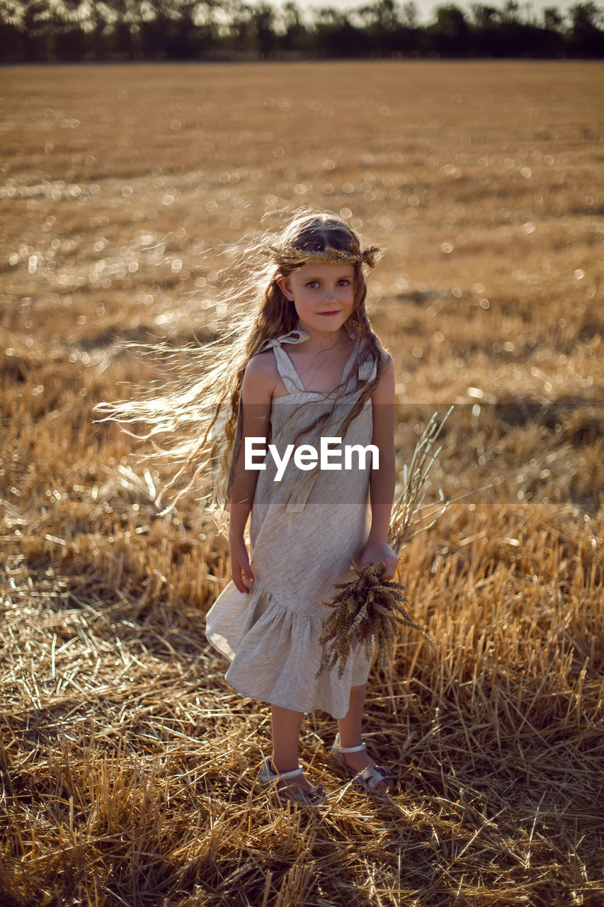 Girl child in a dress and a wreath on her head stands on a mown field of wheat at sunset in summer