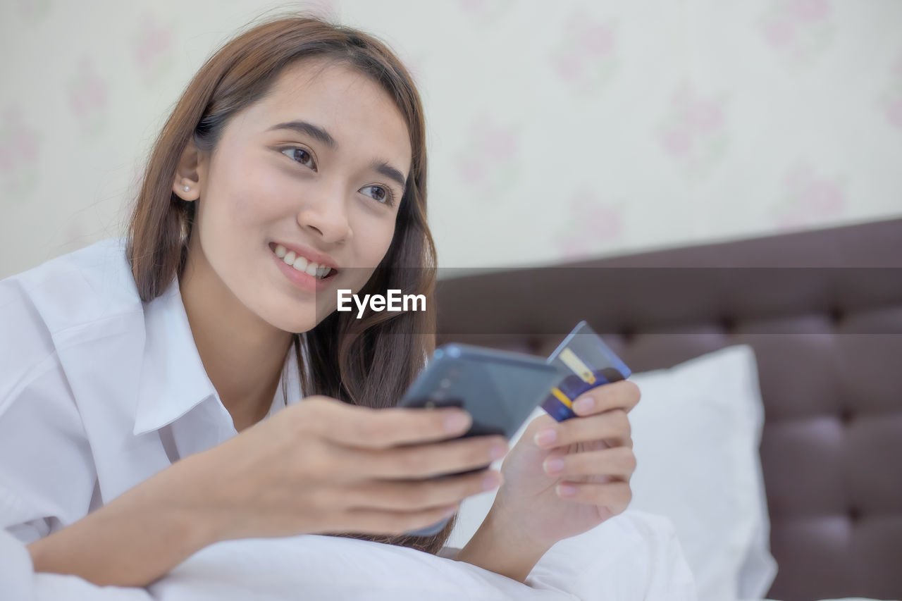 PORTRAIT OF SMILING YOUNG WOMAN USING SMART PHONE WHILE STANDING ON BED