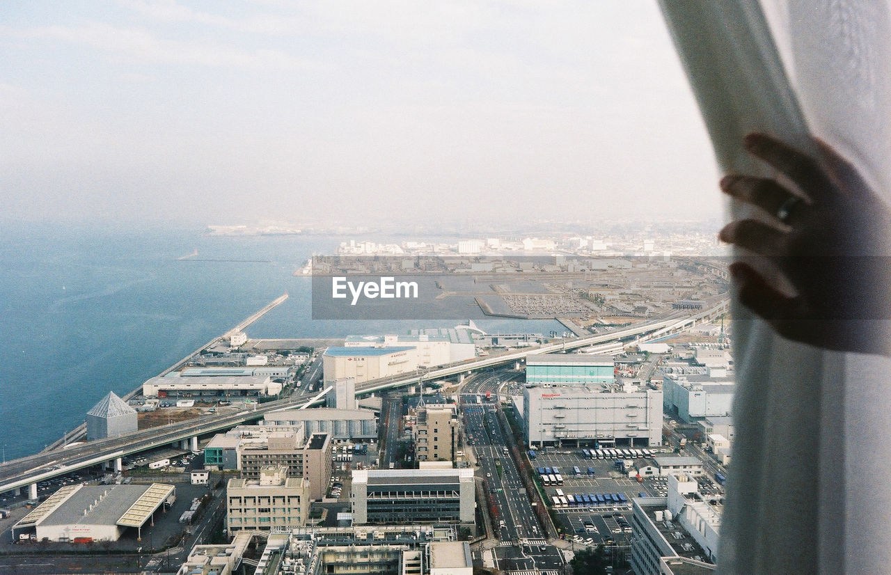 Cropped image of person touching curtain in city against sky