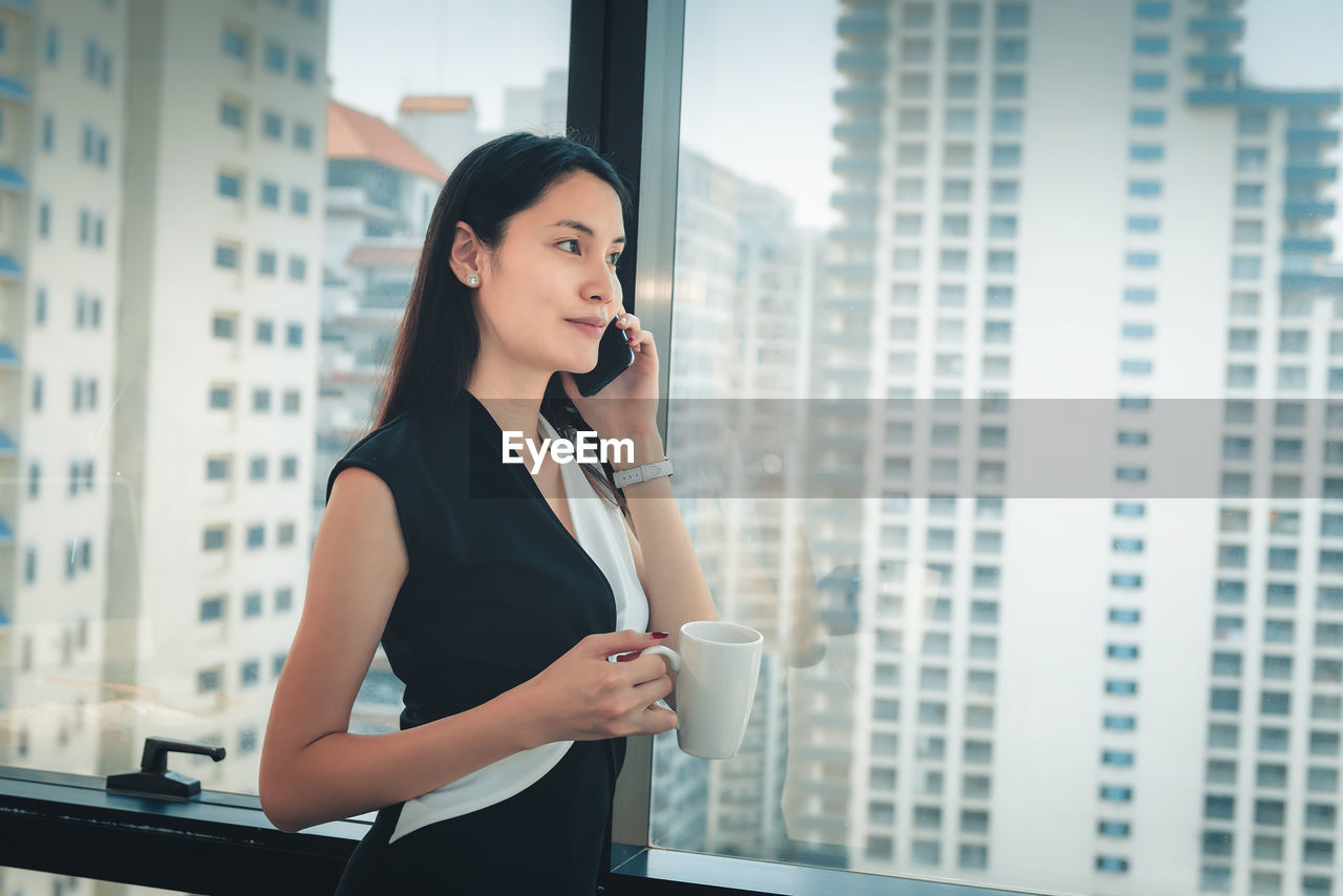 Businesswoman holding coffee cup and using phone while standing by window