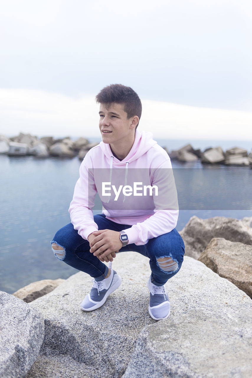 Young man looking away while crouching on rock against sea