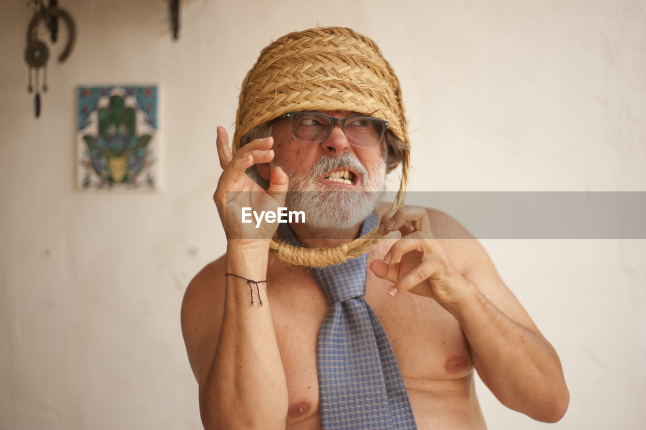 Shirtless senior man with basket on head standing against wall at home