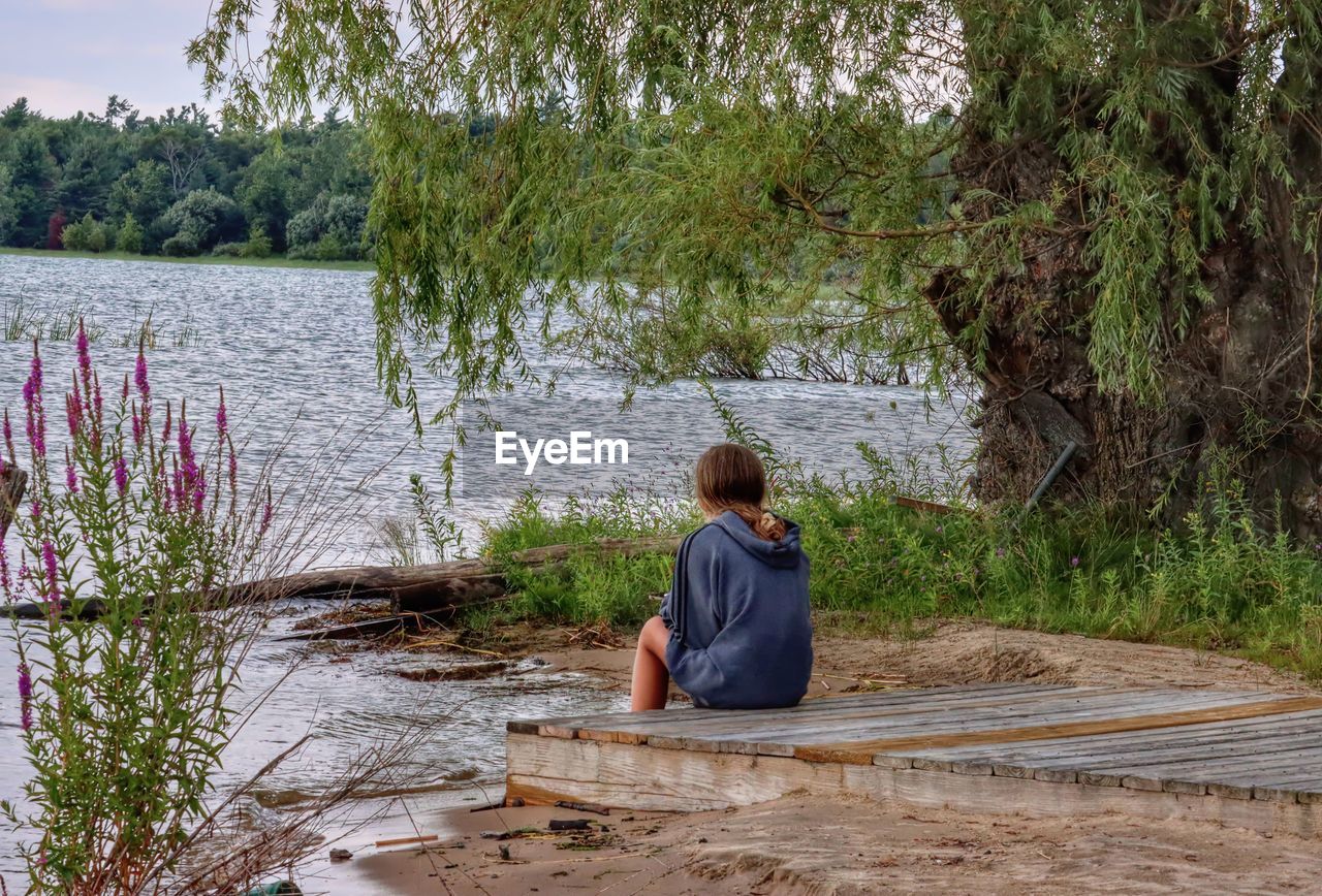 Rear view of young lady sitting by lake against trees