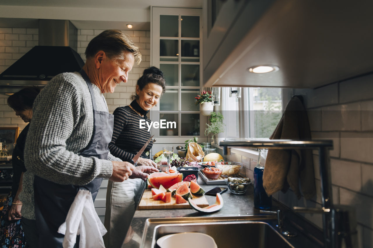 Smiling senior male and female friends cutting fruits at kitchen counter