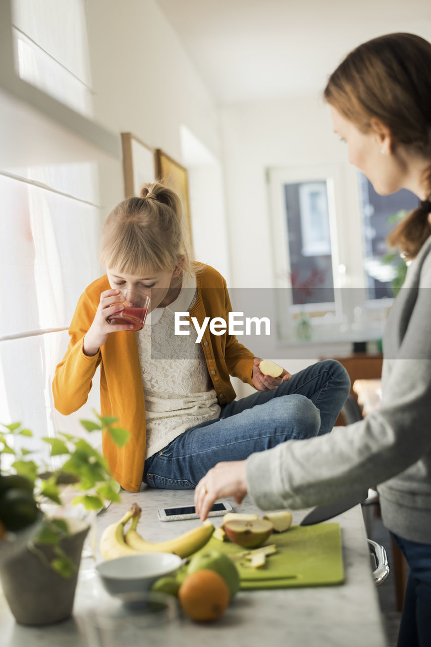 Woman cutting fruits with girl drinking juice on kitchen counter at home