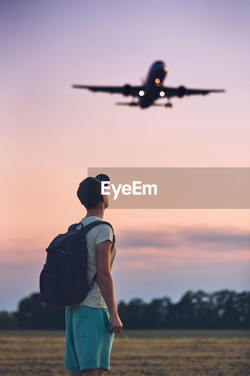 Side view of man with backpack looking at airplane against sky during sunset