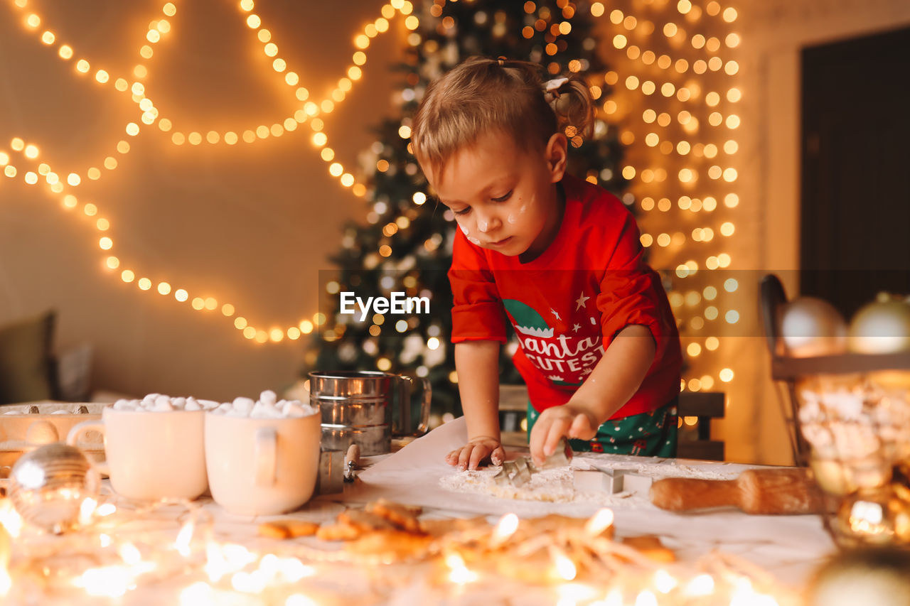 A little girl in red pajamas cooks and eats christmas cookies in the decorated kitchen of the house