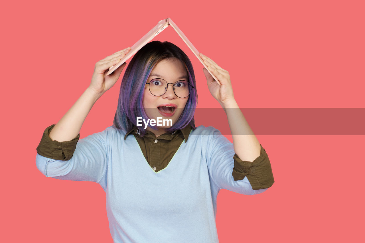 one person, studio shot, emotion, portrait, colored background, eyeglasses, women, pink, indoors, glasses, waist up, fun, facial expression, adult, happiness, front view, red, looking at camera, casual clothing, clothing, young adult, child, shouting, smiling, positive emotion, female, arm, person, cut out, human face, copy space, standing, humor, gesturing, childhood, finger, hand, hairstyle, mouth open, cheerful, arms raised, negative emotion, t-shirt, frustration