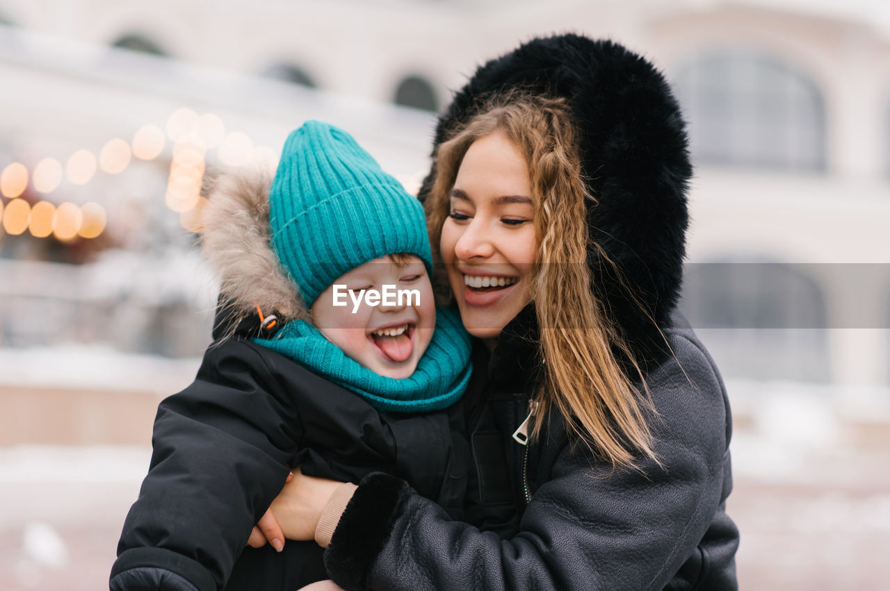 Happy woman is holding a child in a blue hat and a black coat. joyful boy is smiling and making