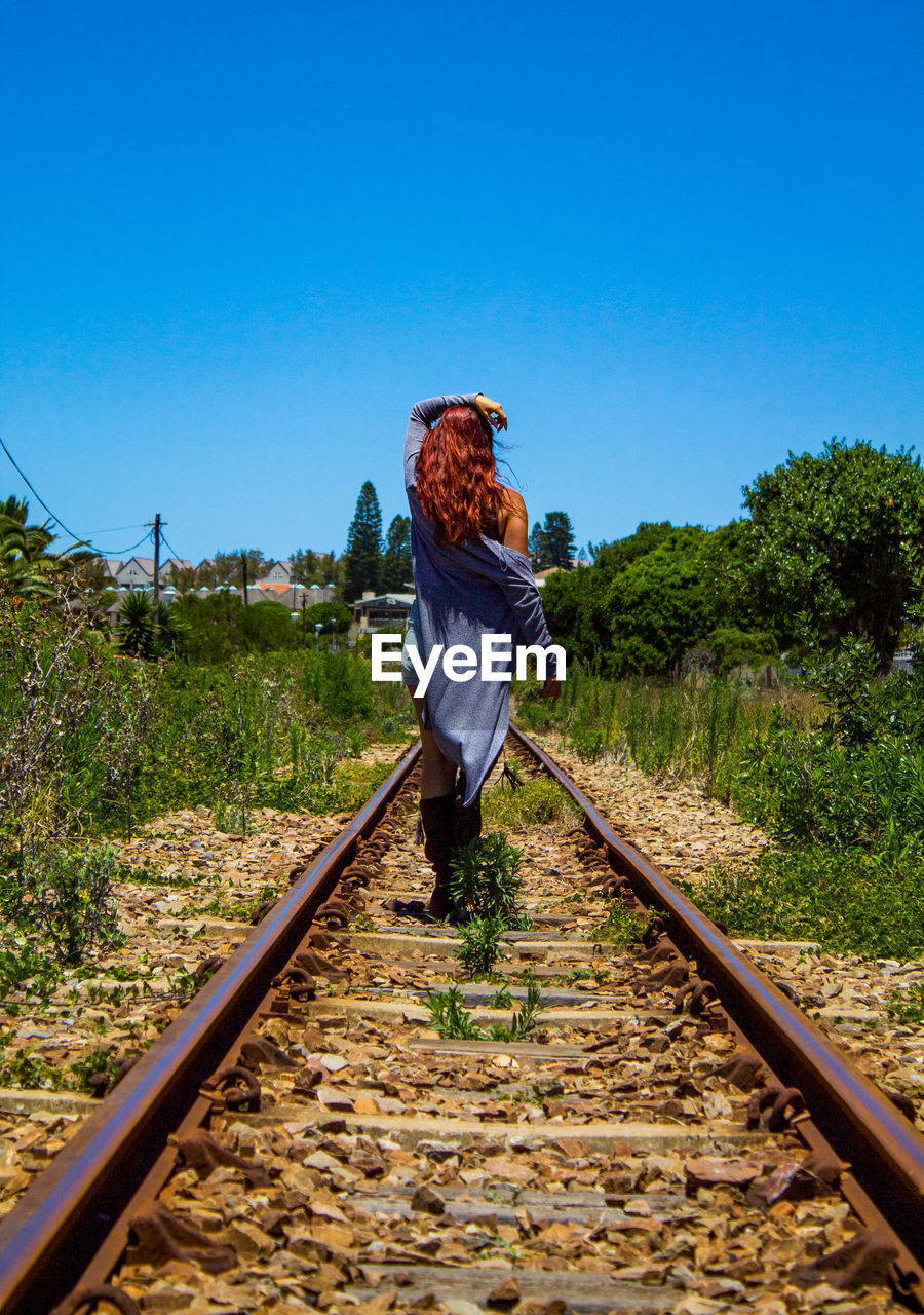 REAR VIEW OF WOMAN WALKING ON RAILROAD TRACKS AGAINST SKY