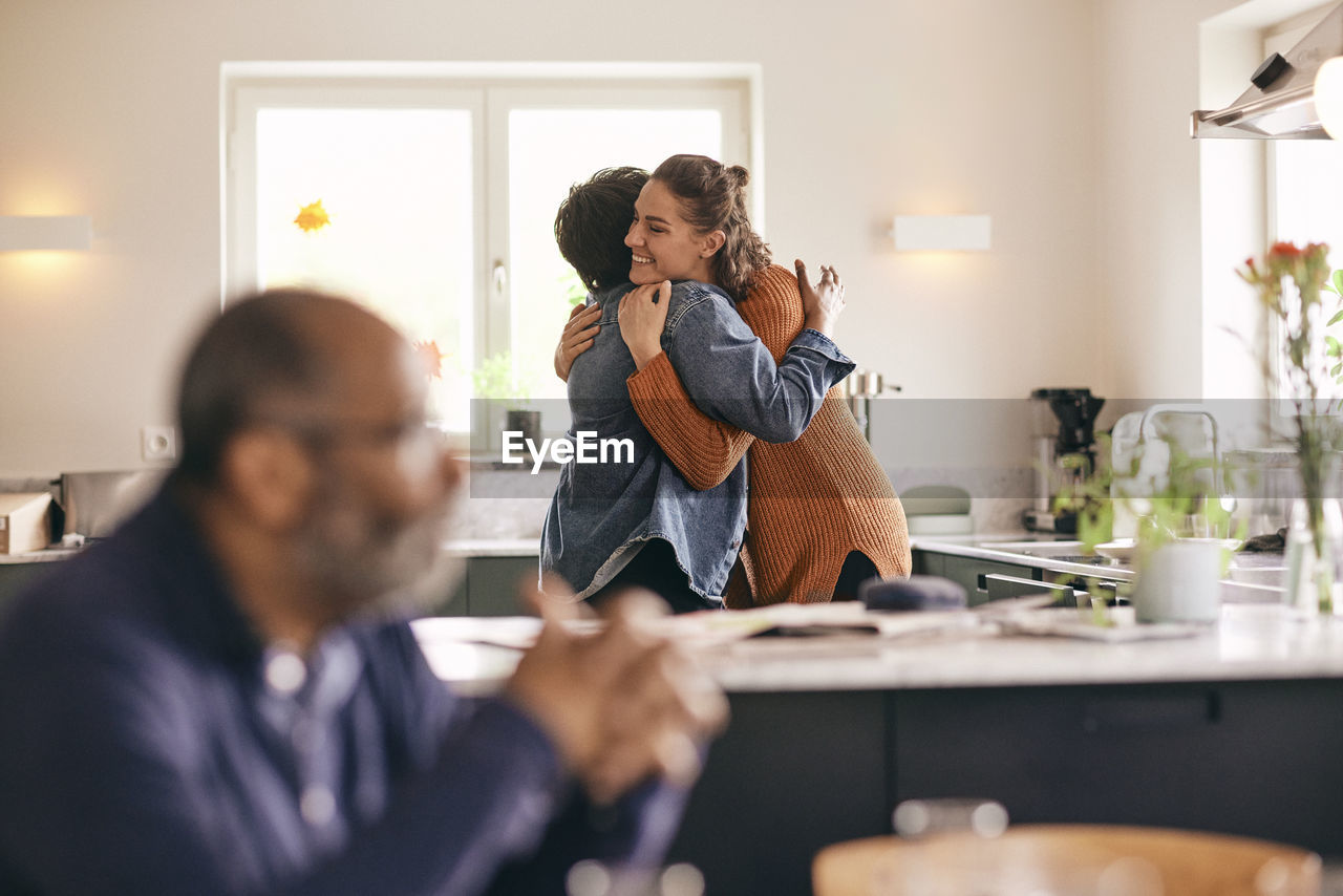 Smiling woman embracing mother-in-law while standing in kitchen at home