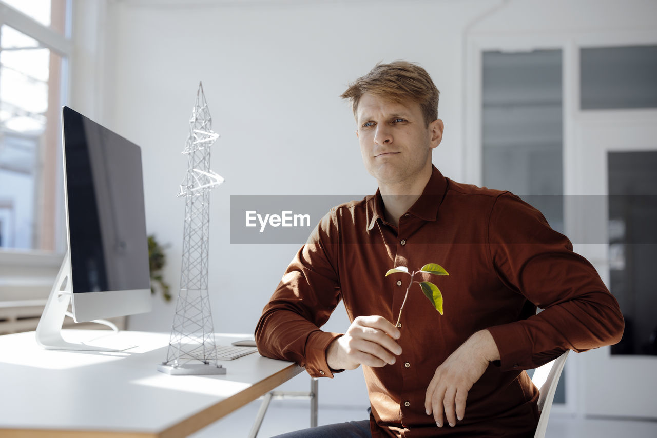 Thoughtful engineer holding leaf sitting by electricity pylon model at desk in office