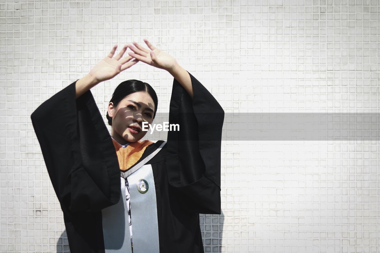 Portrait of young woman wearing graduation gown shielding eyes against wall