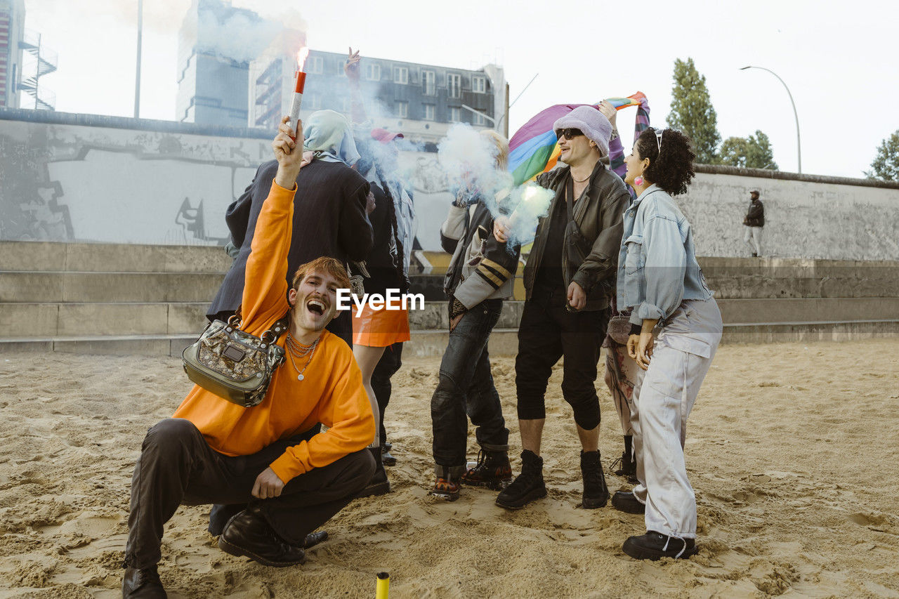 Gay man holding distress flare while crouching near non-binary friends on sand at park