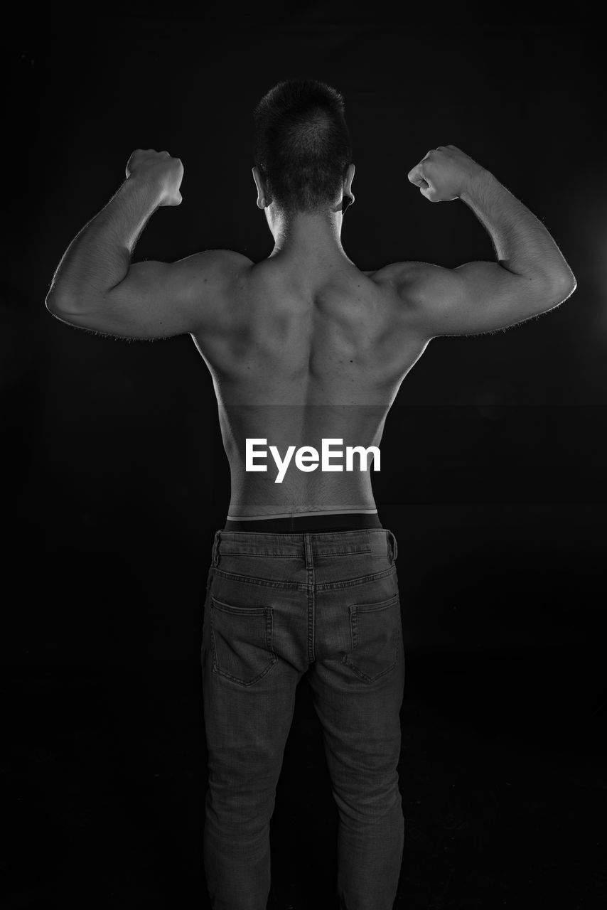 Rear view of shirtless man flexing muscles against black background