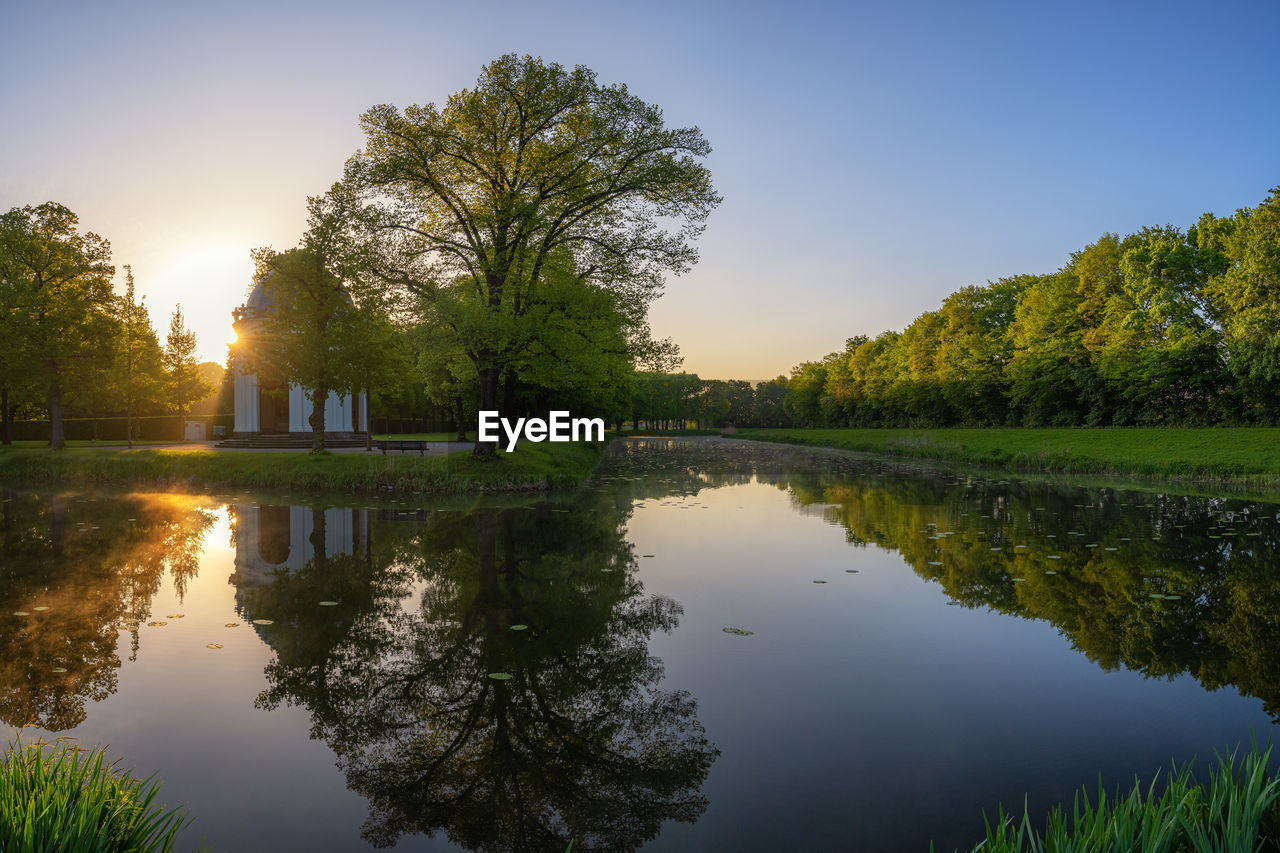 reflection, water, nature, tree, plant, lake, sky, beauty in nature, autumn, tranquility, environment, morning, landscape, scenics - nature, sun, leaf, tranquil scene, architecture, summer, travel destinations, twilight, outdoors, reflection lake, no people, blue, sunrise, sunlight, travel, grass, body of water, land, tourism, multi colored, social issues, clear sky, reservoir, idyllic, building, rural scene, forest, waterway, activity, built structure, non-urban scene, green