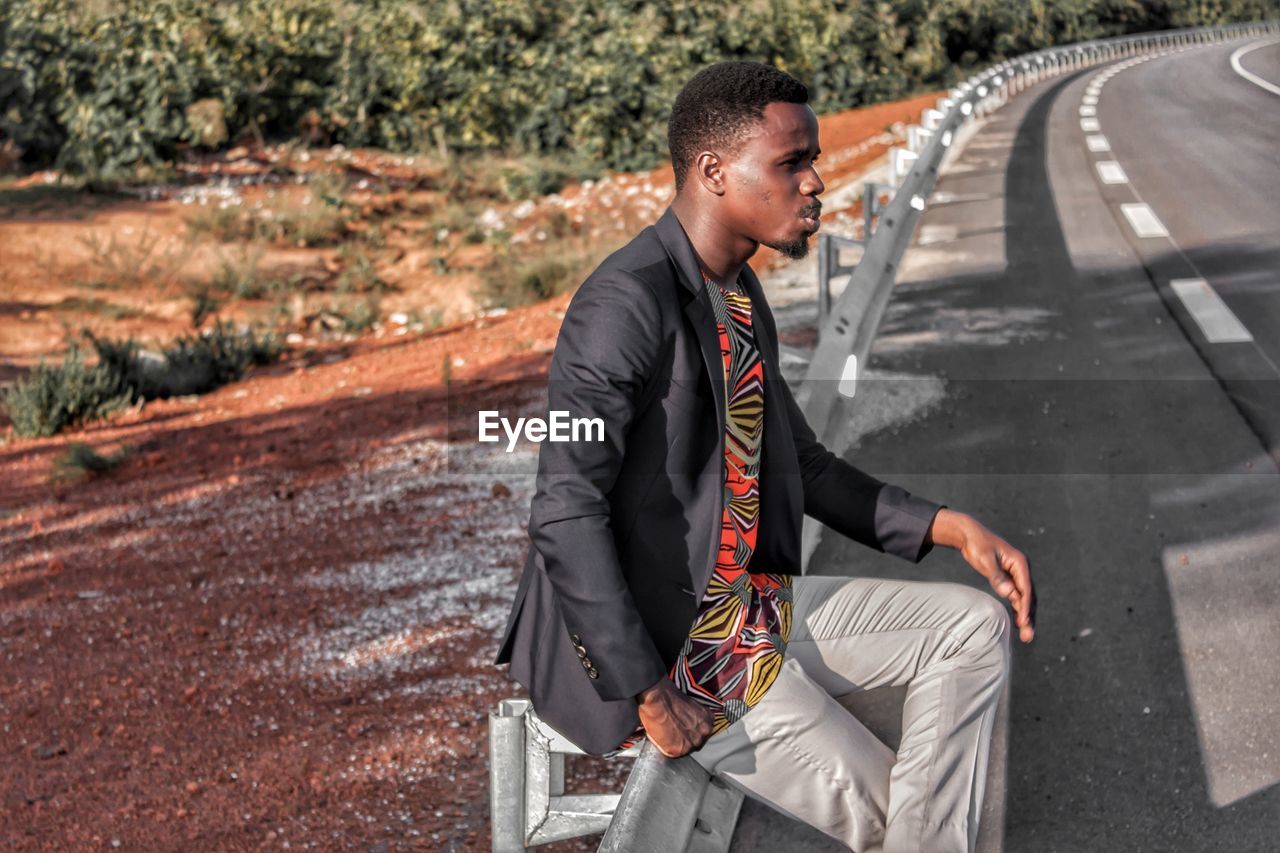 Side view of young man sitting on railing by road against sky