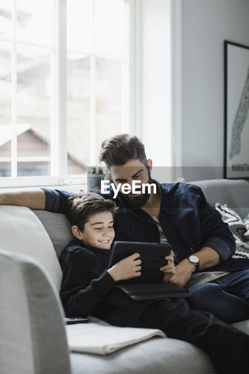 Happy father looking at boy using digital tablet while sitting on sofa in living room