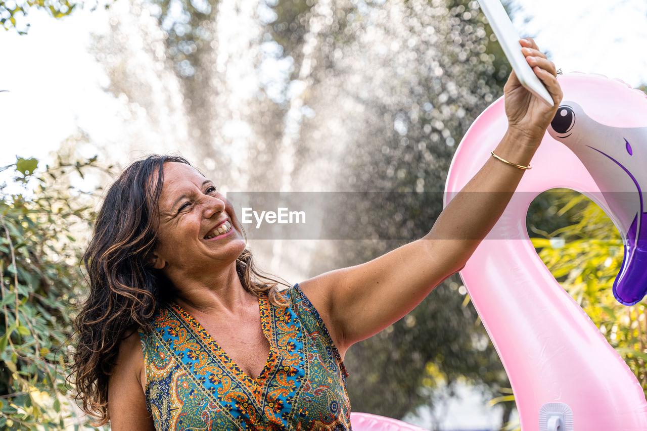 Beautiful smiling middle aged woman taking a selfie sitting on a pink flamingo shaped inflatable toy