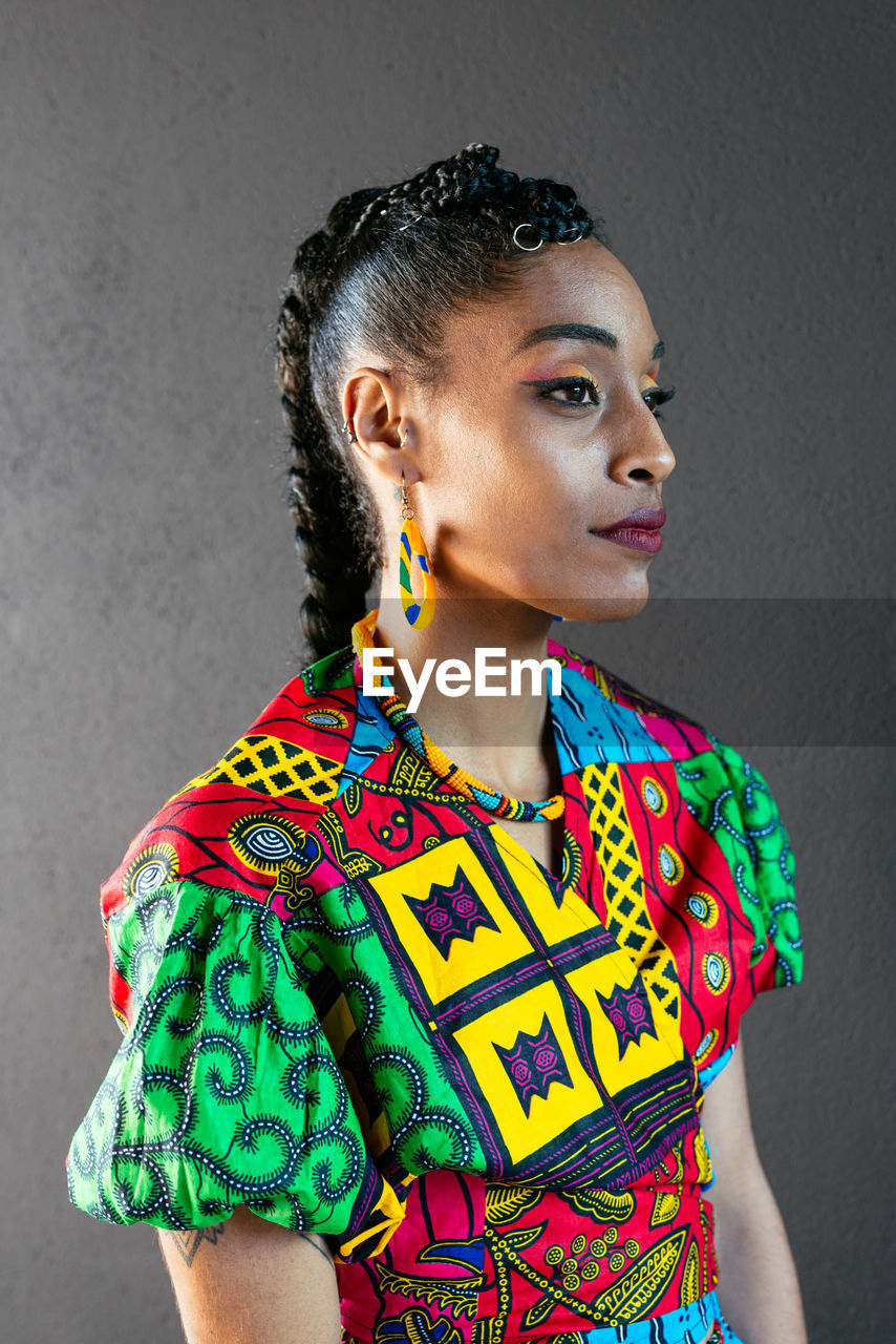 Ethnic female in multicolored ornamental garment with braided hair looking away against gray wall