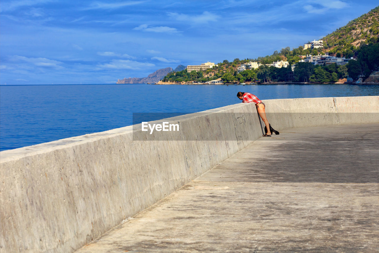 A lady in a beach suit watches the sea fish leaning over the concrete pier of the sea bay.