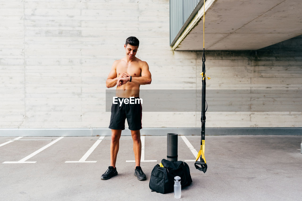 Content shirtless sportsman standing on street and checking pulse on fitness tracker after active workout in city