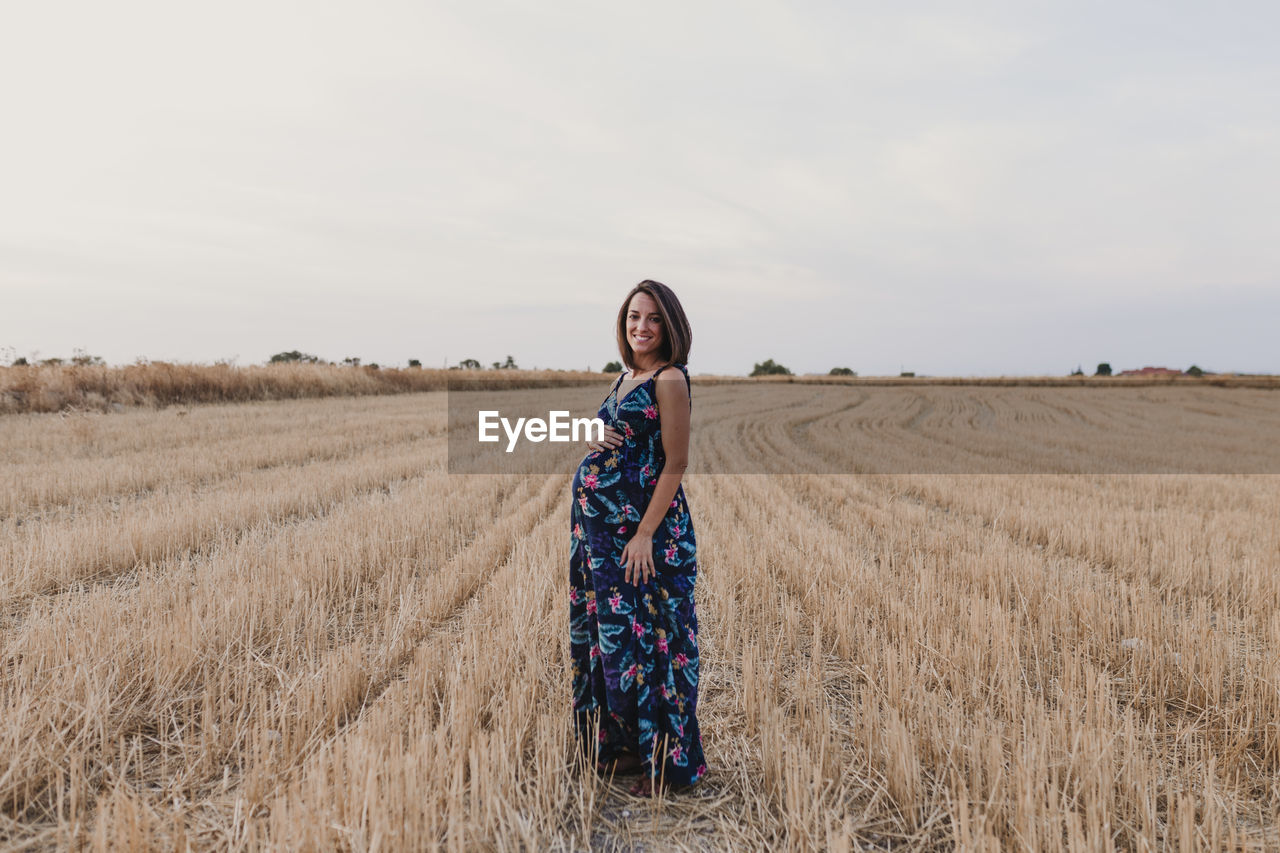 Portrait of smiling pregnant woman with hand on stomach standing on field