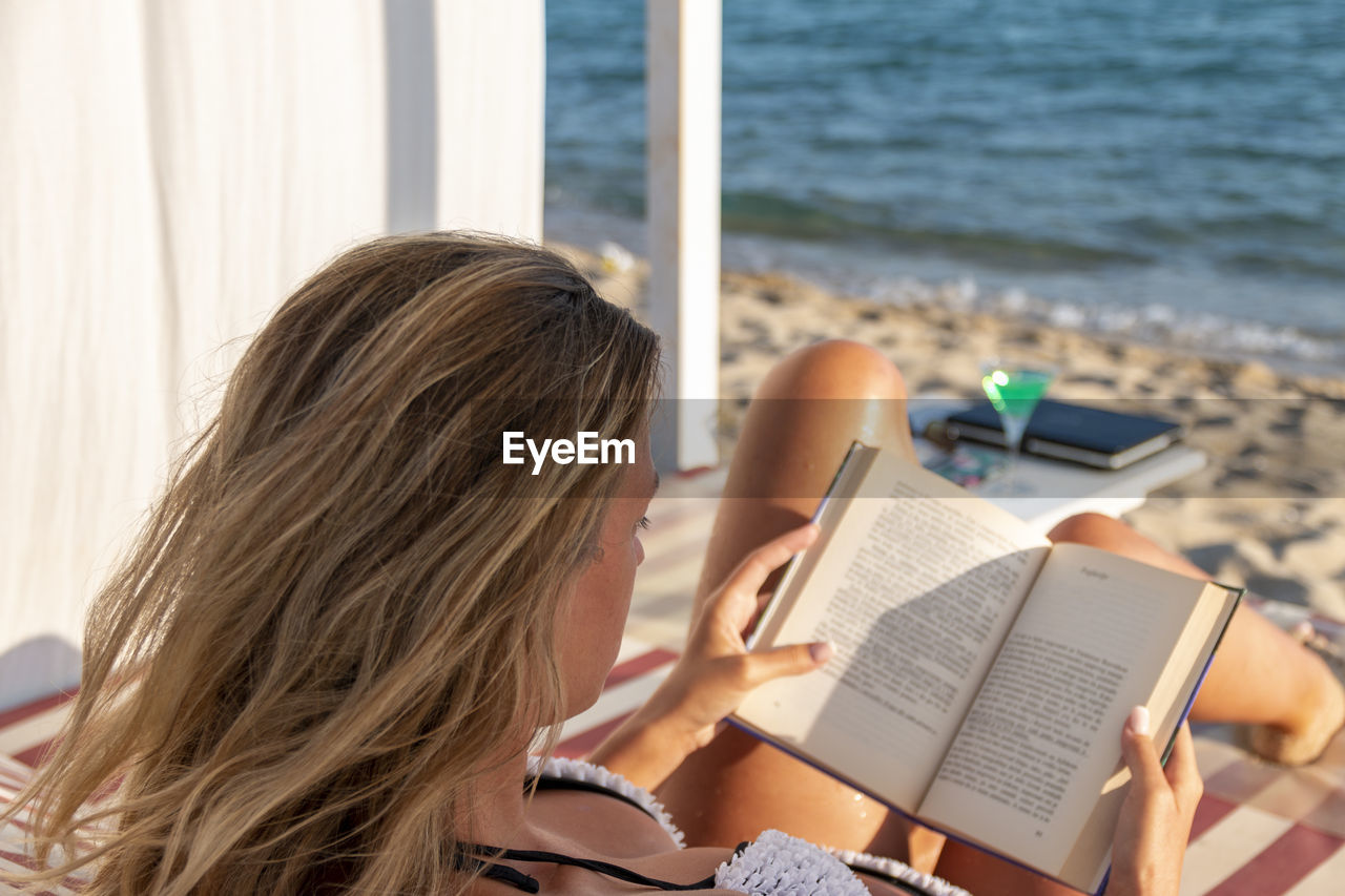 Young blonde woman reading a book and relaxing on a daybed iat the beach