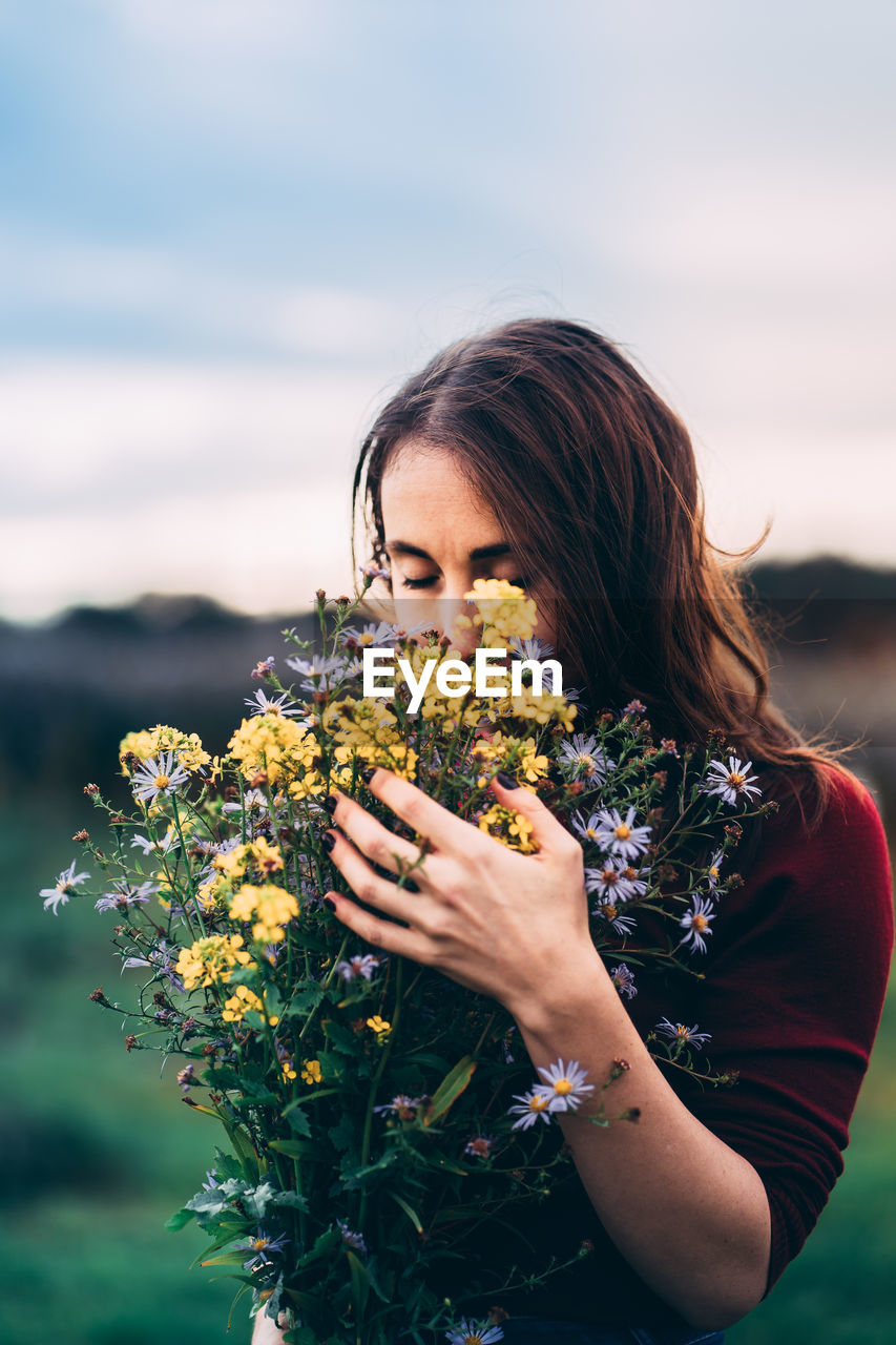 Woman with long brown hair in red sweater thoughtfully holding bouquet of wild flowers in the nature