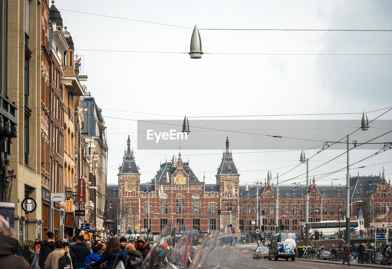 People on road against amsterdam centraal railway station and sky in city