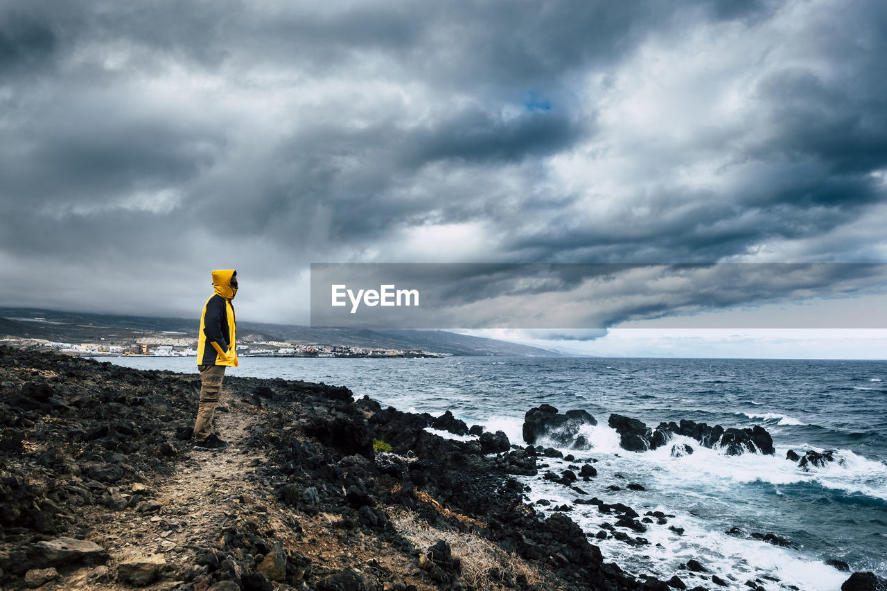 Side view of man standing at beach against cloudy sky