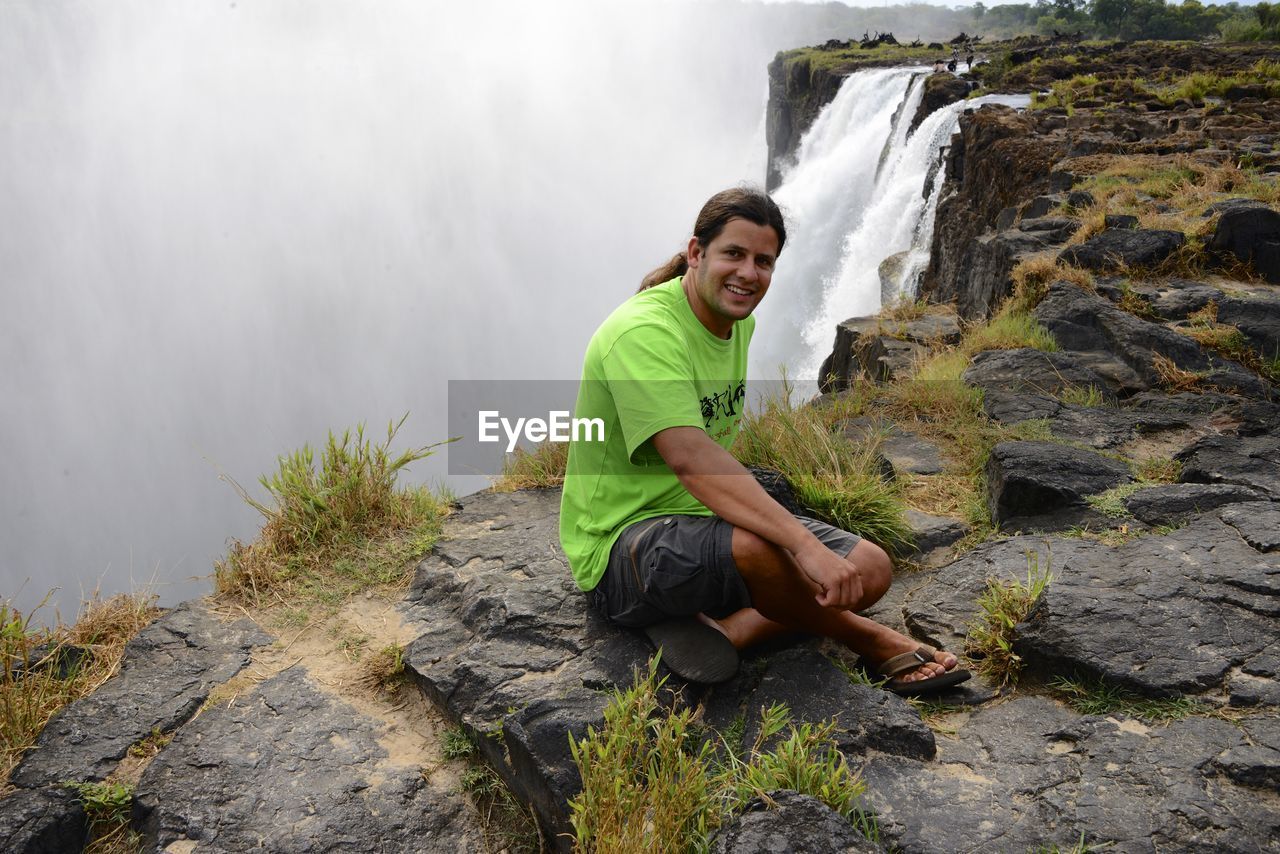 Man sitting on cliff against waterfall