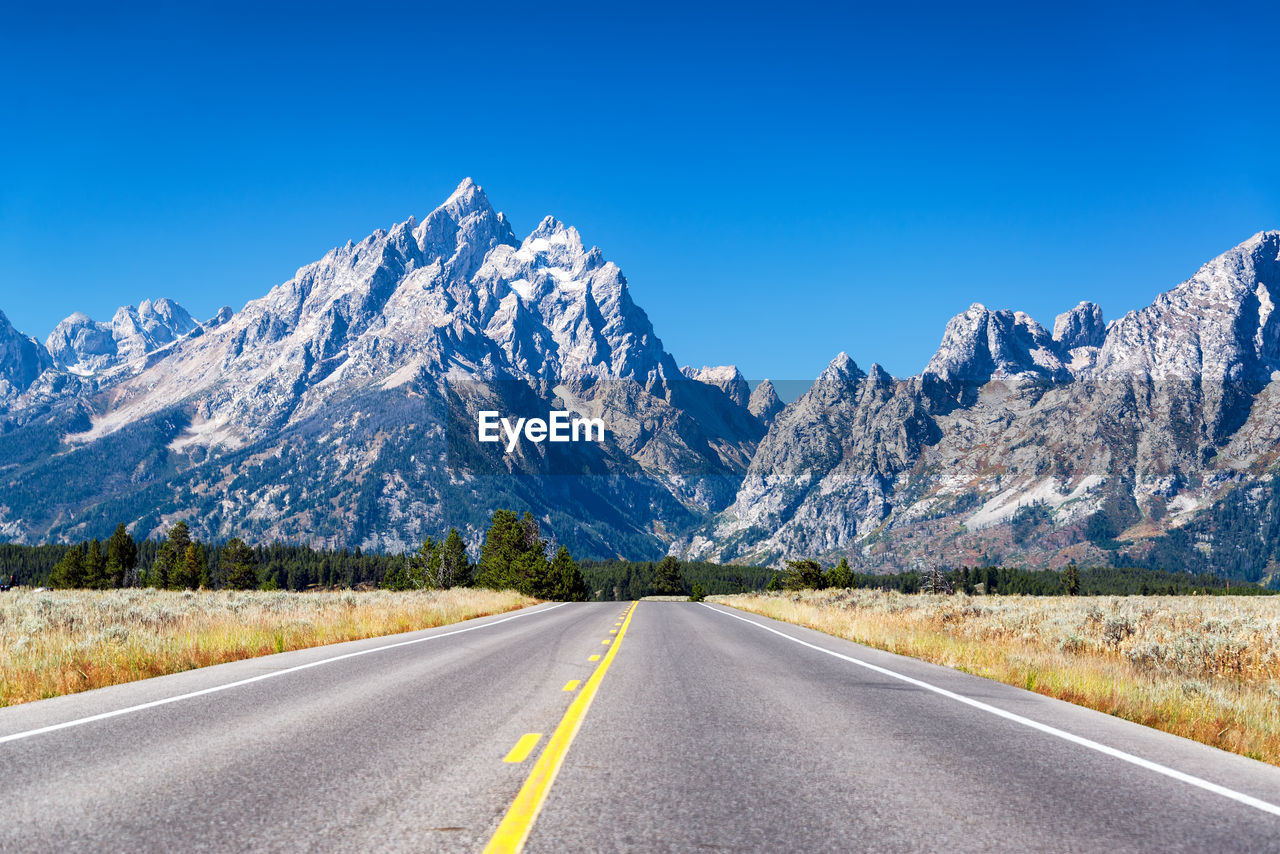 Road amidst field leading towards snowcapped mountains against clear blue sky