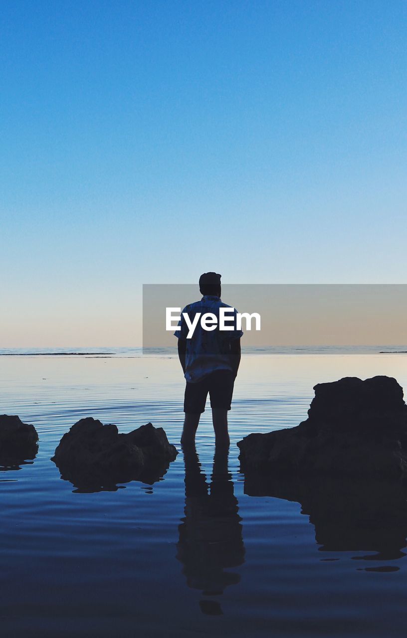 REAR VIEW OF SILHOUETTE MAN STANDING AT BEACH AGAINST SKY