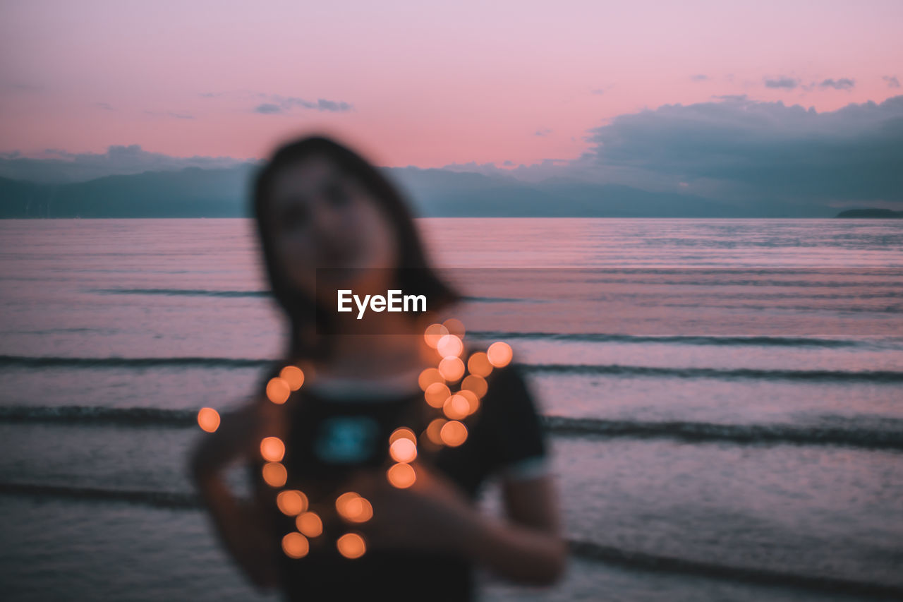 Defocused image of woman wearing illuminated lights at beach against sky during sunset